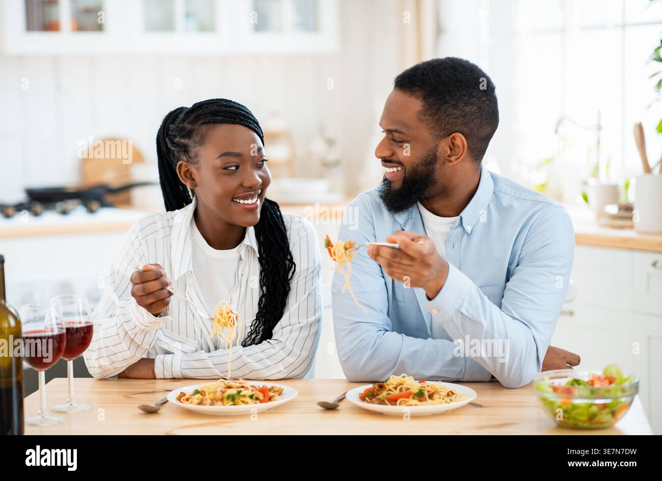 Ein schwarzes Paar, das sich ansieht, wie er Pasta isst und zu Hause in der Küche sitzt Stockfoto