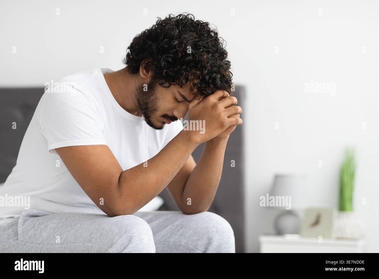 Gestresster bärtiger indischer Mann mit lockigem Haar, der die Stirn auf geballten Fäusten lehnt und im Schlafzimmer zu Hause auf dem Bett sitzt Stockfoto