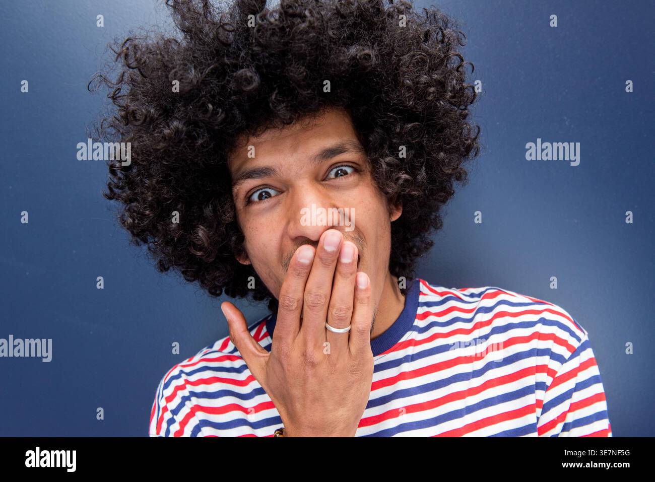 Überraschter afroamerikanischer Mann mit Hand, die den Mund bedeckt, steht an der blauen Wand Stockfoto