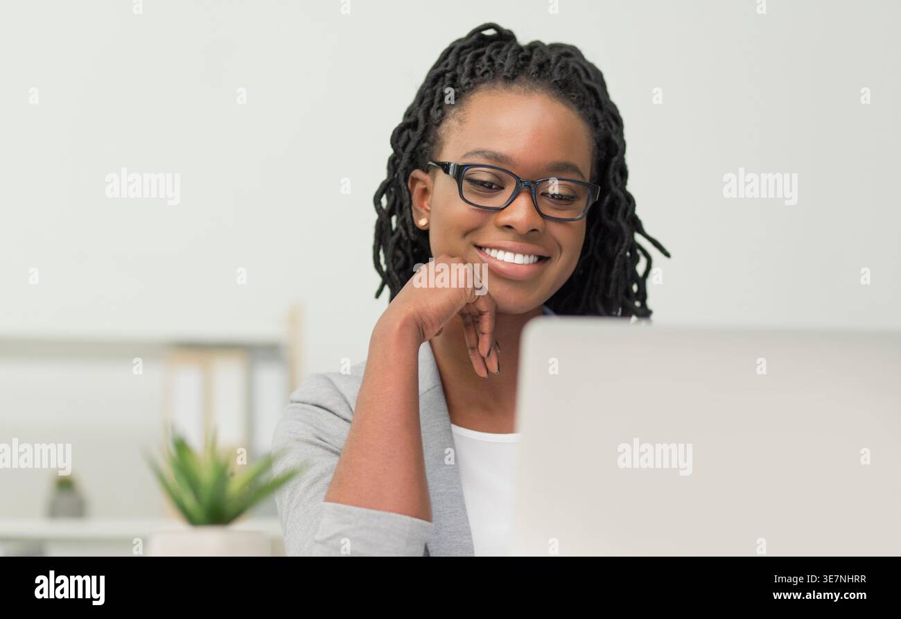 Lächelnde schwarze Geschäftsfrau mit Twists, die grauen Mantel und Brille trägt und auf den Laptop blickt, während sie am Tisch im Büro sitzt Stockfoto