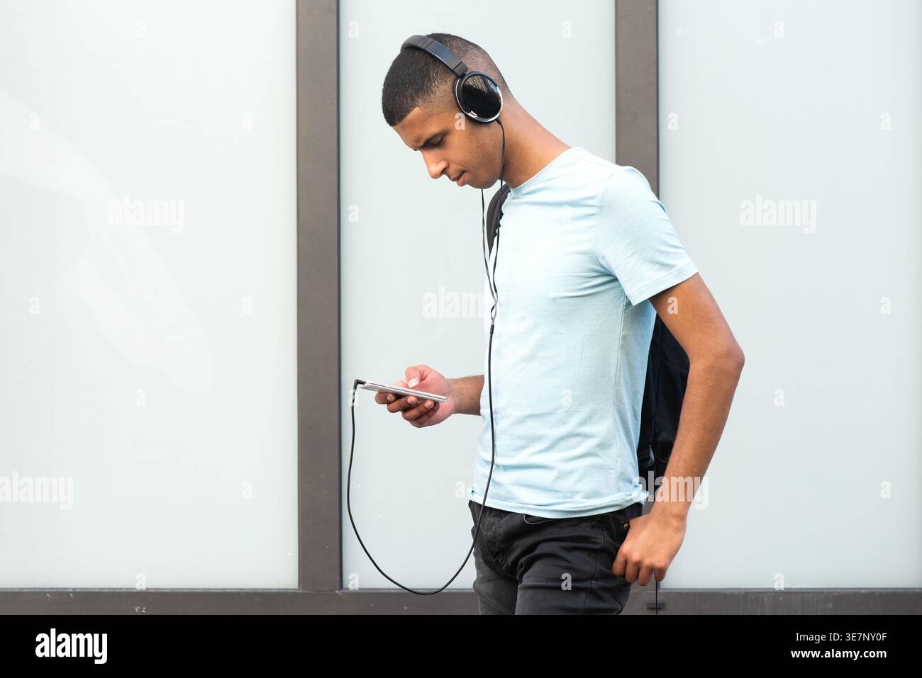 Ernster arabischer männlicher Teenager in Kopfhörern mit Smartphone, das tagsüber mit dem Daumen in der Tasche gegen die Wand steht Stockfoto