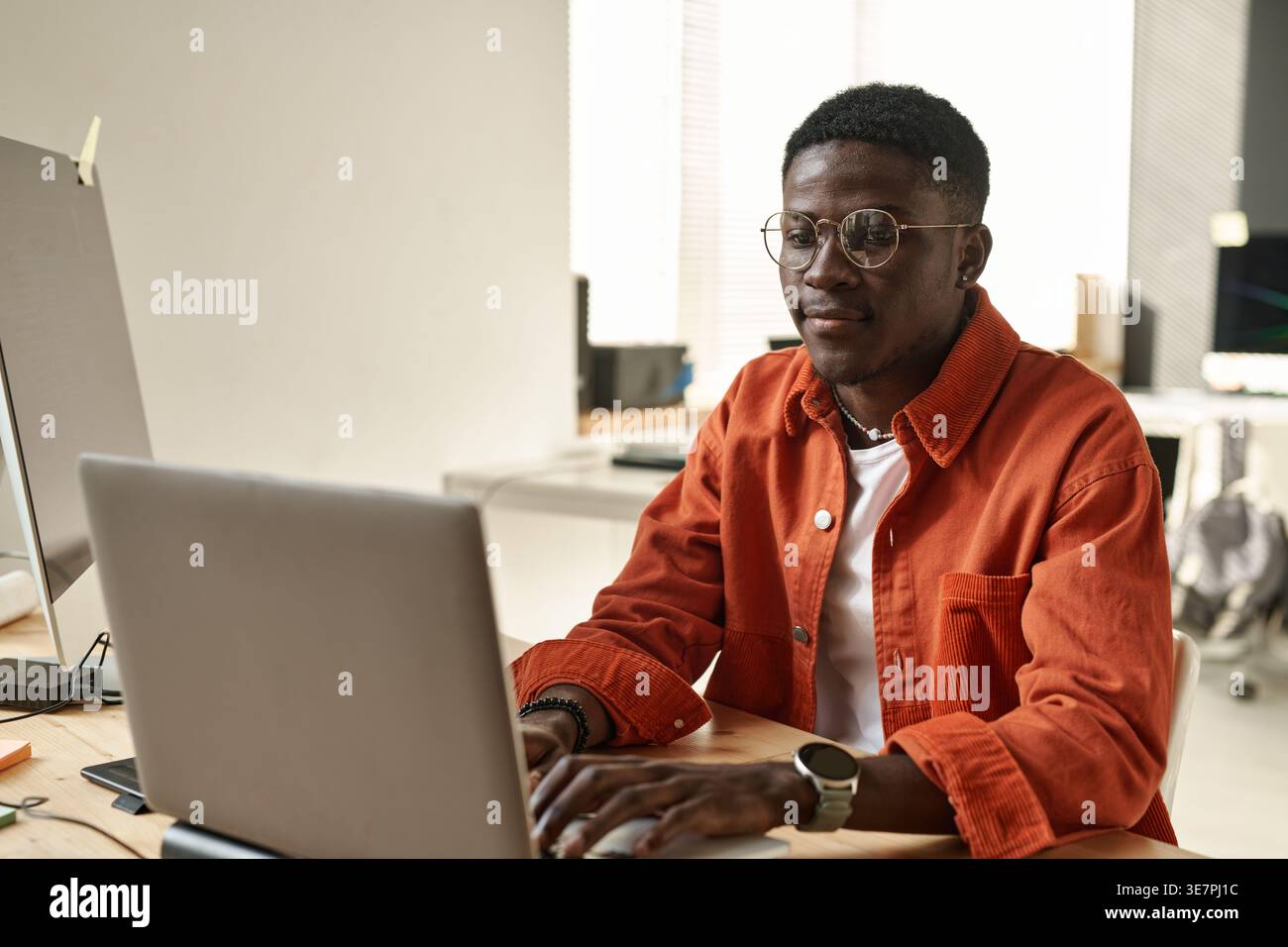 Schwarzer, männlicher Software-Entwickler mit Brille, der am Laptop arbeitet und am Tisch im Büro sitzt Stockfoto