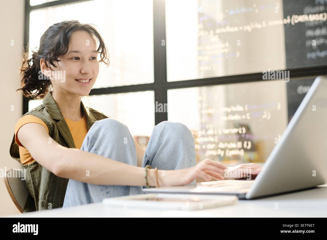 Lächelnde gemischte Schülerin, die im Klassenzimmer mit den Füßen unter den Beinen sitzt Stockfoto