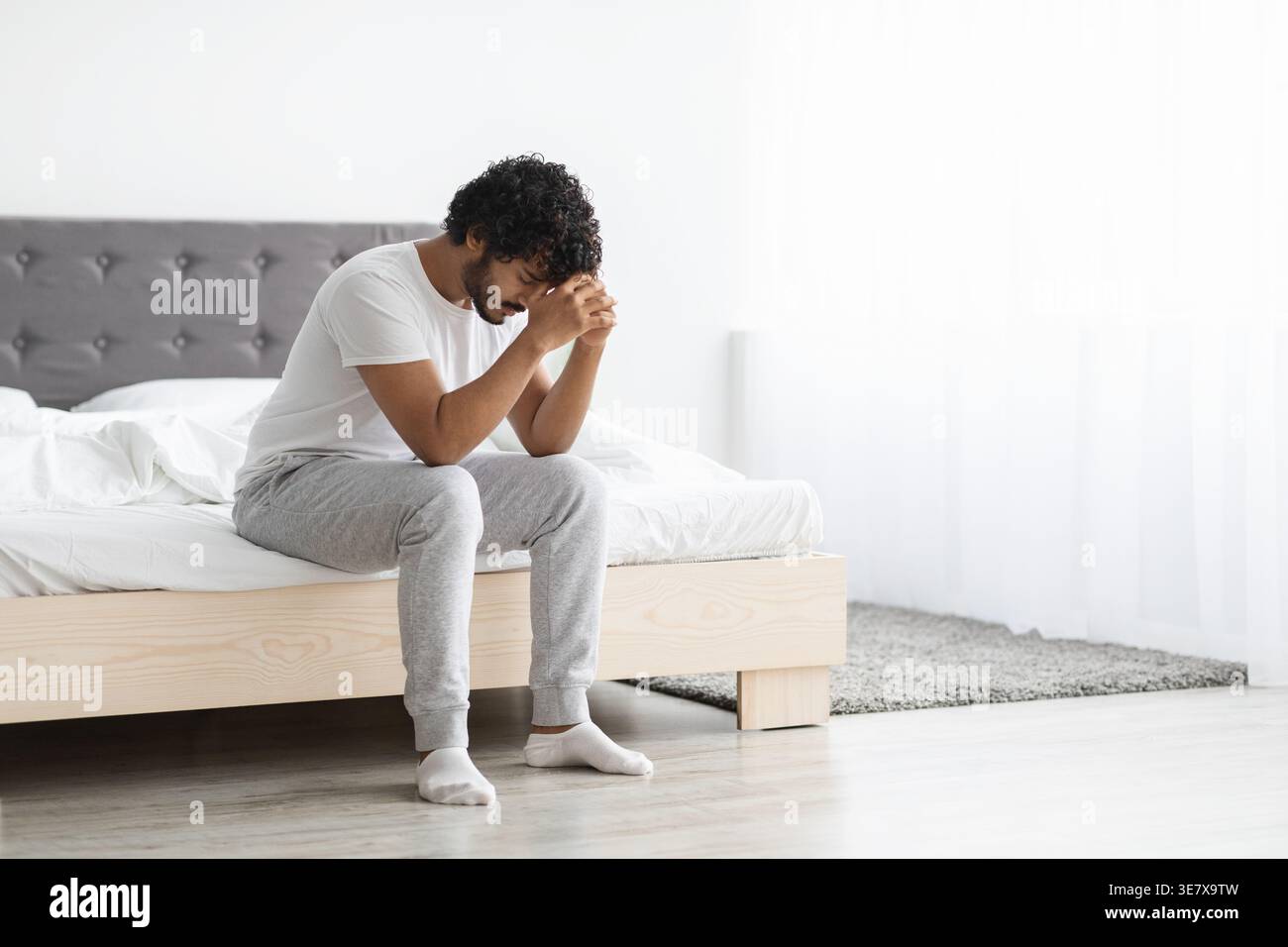 Gestresster bärtiger indischer Mann mit lockigem Haar, der die Stirn auf geballten Fäusten lehnt und im Schlafzimmer zu Hause auf dem Bett sitzt Stockfoto