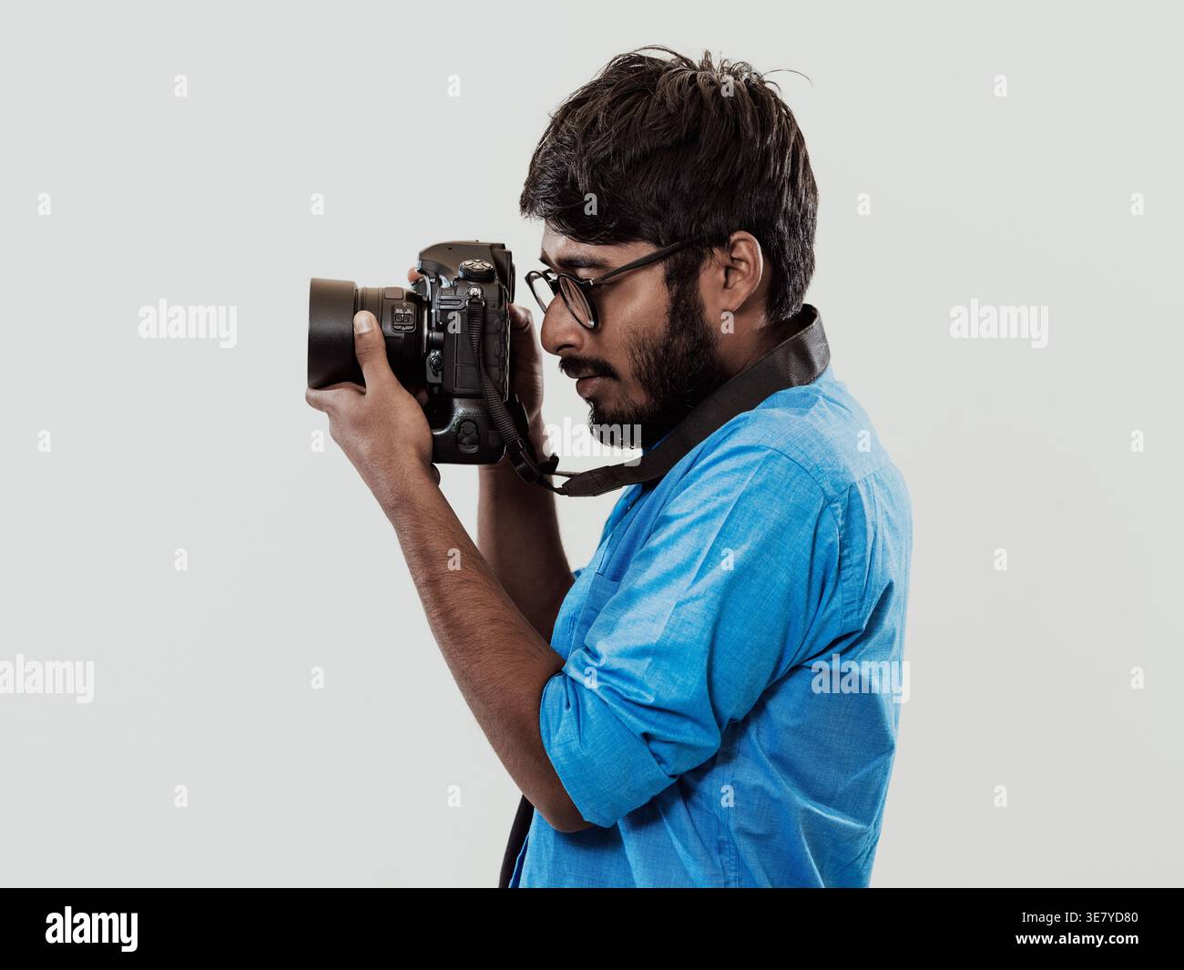 Lächelnder indischer Mann in blauen langen Ärmeln mit Brille, der Fotos mit einer DSLR-Kamera macht, während er im Studio vor weißem Hintergrund steht Stockfoto