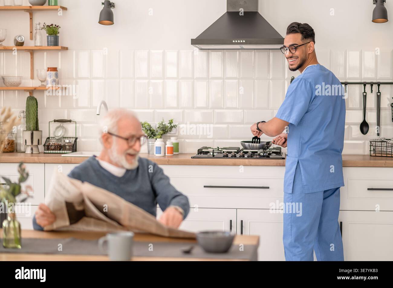 Lächelnder arabischer männlicher Pfleger in Brille kocht im Stehen, während er die Zeitung ansieht, wie ein älterer Mann zu Hause am Tisch sitzt Stockfoto