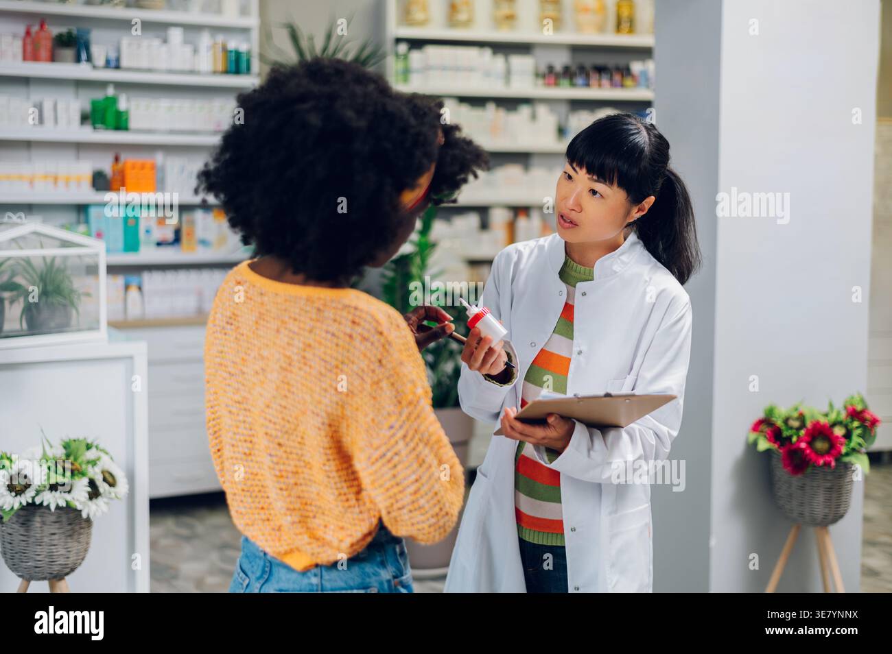 Japanische Apothekerin mit Salbenspender und Klemmbrett, die mit schwarzer Patientin und Kundin in der Apotheke spricht Stockfoto