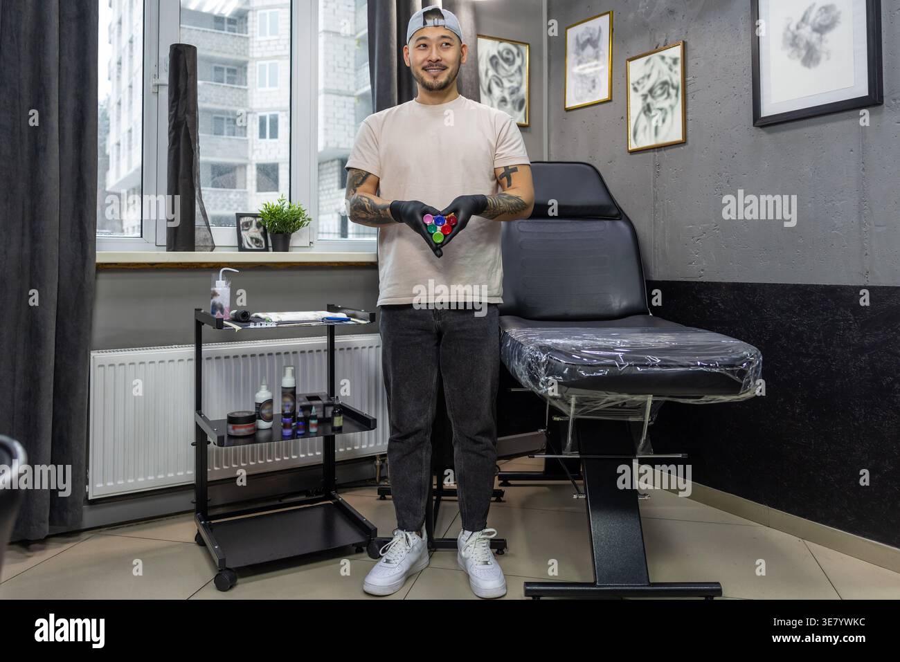 Lächelnder asiatischer Tätowierer mit Mütze und Latexhandschuhen, der verschiedene Tinten hält, bereitet sich auf die Tätowierung im Studio vor Stockfoto