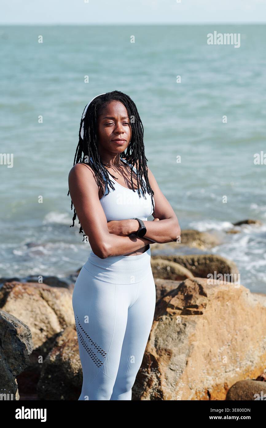 Schwarze Frau mit Dreadlocks im Fitness-Outfit mit überkreuzten Armen an sonnigen Tagen am Meer Stockfoto