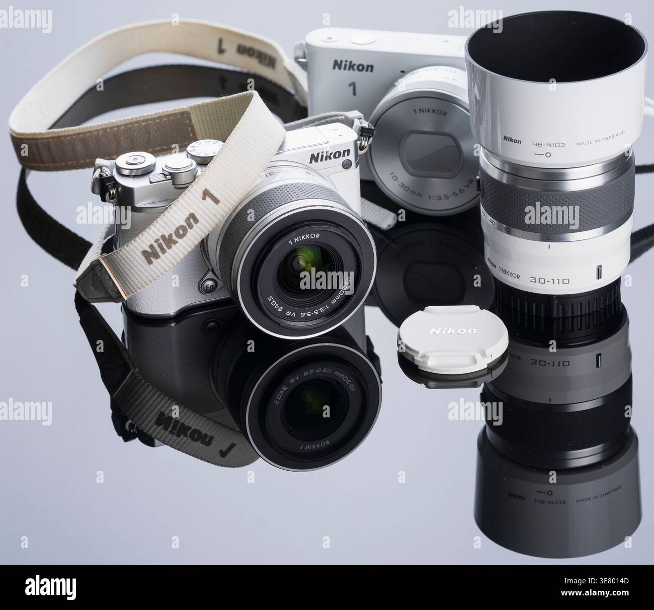 Eine weiße Nikon 1 spiegellose Kamera mit 1 NIKKOR Objektiv, Objektivblende und Objektivkappe wird auf einer glänzend schwarzen Oberfläche angezeigt Stockfoto
