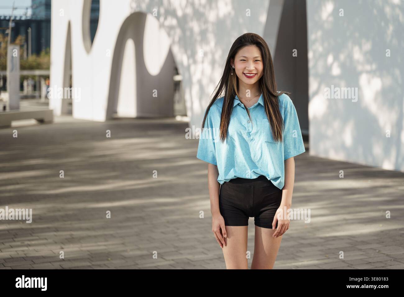 Lächelnde asiatische Frau, die am sonnigen Tag auf der Straße in der Nähe des Gebäudes steht Stockfoto