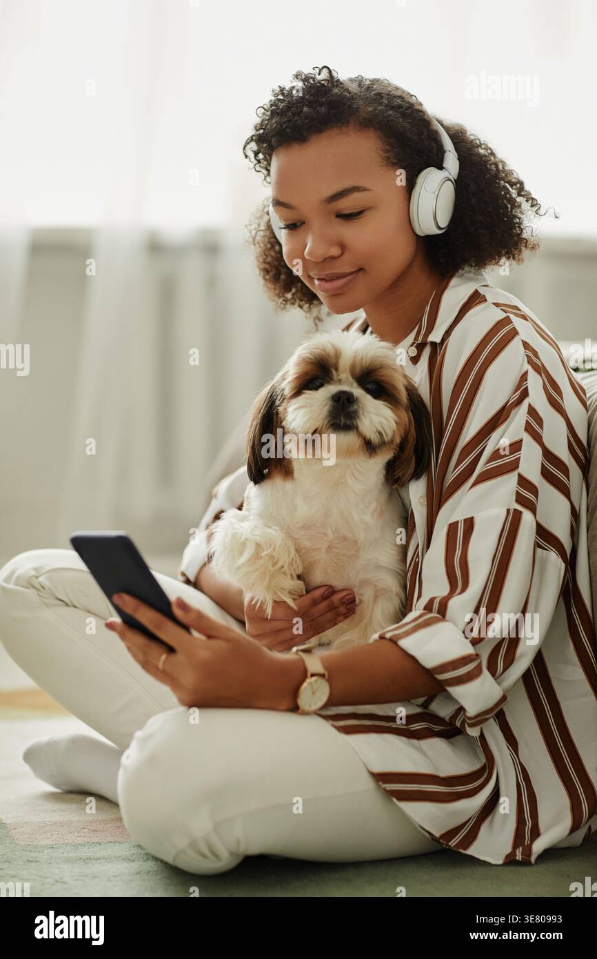 Gemischte Frau mit lockigem Afro, die Kopfhörer verwendet, Musik mit smLifestylephone hören, während sie zu Hause einen Hund auf ihrem Schoß streichelt Stockfoto