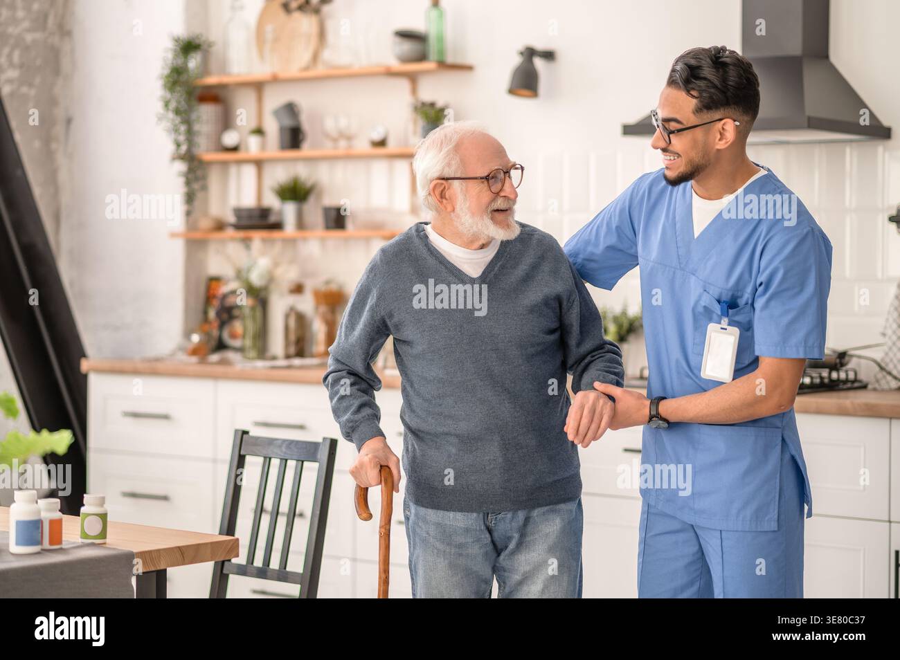 Lächelnder arabischer männlicher Betreuer in Brille, der einen älteren Mann im Stehen zu Hause ansieht Stockfoto
