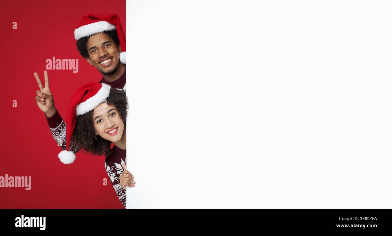 Schwarzes Paar mit Weihnachtsmännern, das hinter weißem Banner vor rotem Hintergrund im Studio guckt Stockfoto