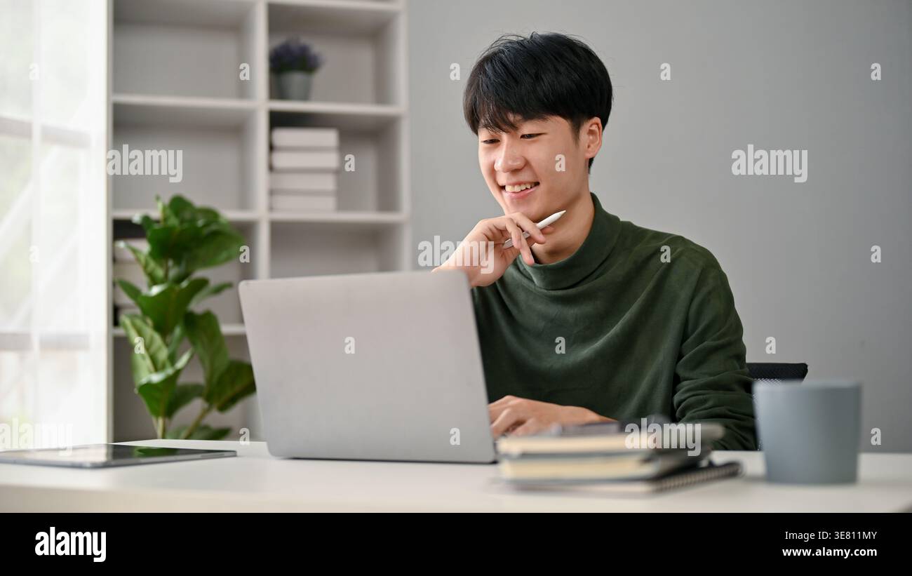 Asiatischer Mann, der am Laptop arbeitet, während er zu Hause am Tisch sitzt Stockfoto