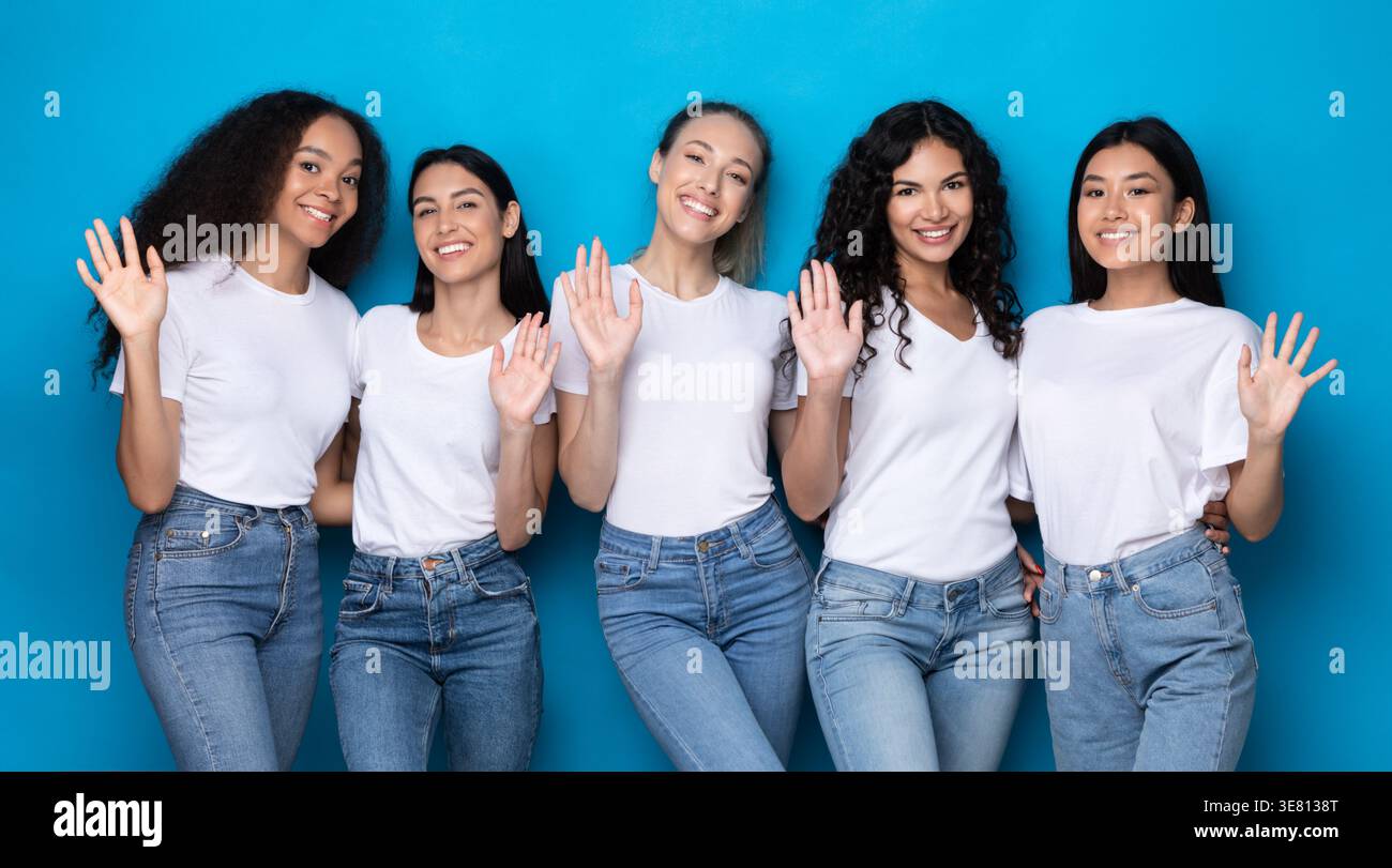 Eine Gruppe von Frauen in weißen Hemden und Jeans, die im Studio vor blauem Hintergrund die Hände winkten Stockfoto
