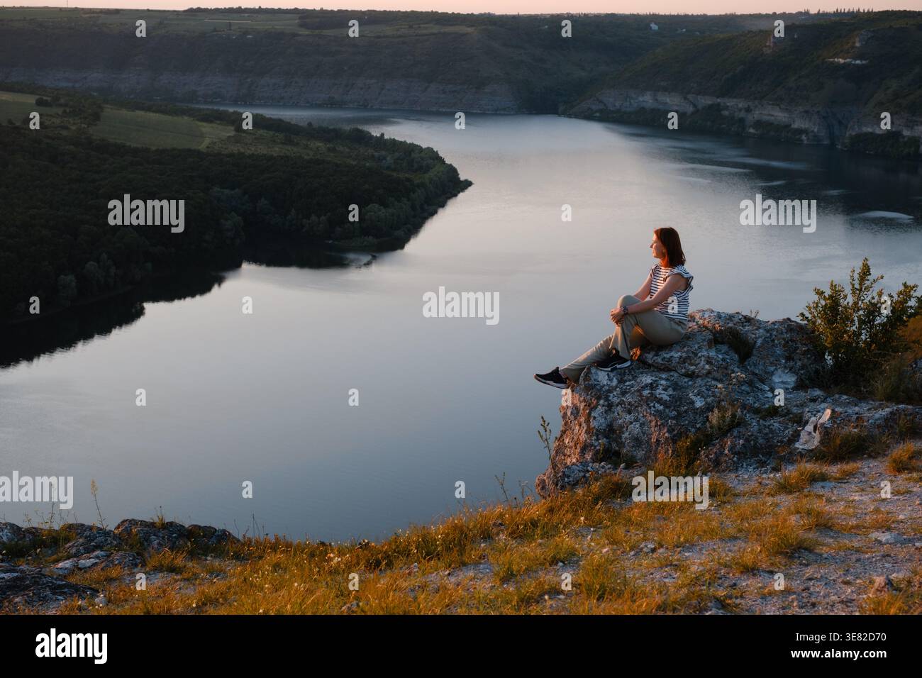 Eine junge Wanderer sitzt an einer felsigen Klippe und genießt bei Sonnenuntergang den malerischen Blick auf den Fluss Canyon. Friedliche Reisende, die sich während der goldenen Stunde in der Natur ausruhen Stockfoto