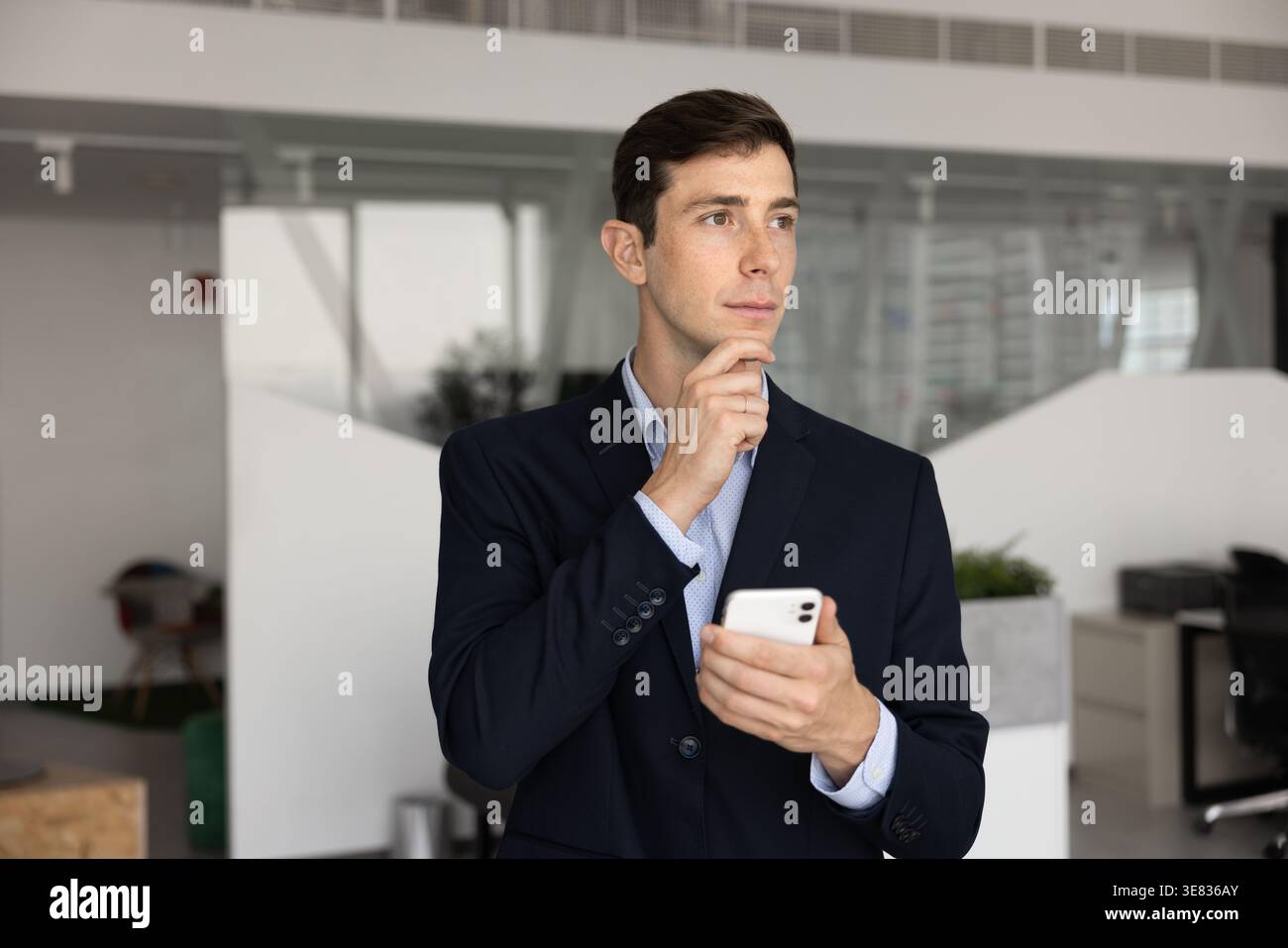 Nachdenklicher Geschäftsmann im Anzug steht im Büro mit Smartphone Stockfoto