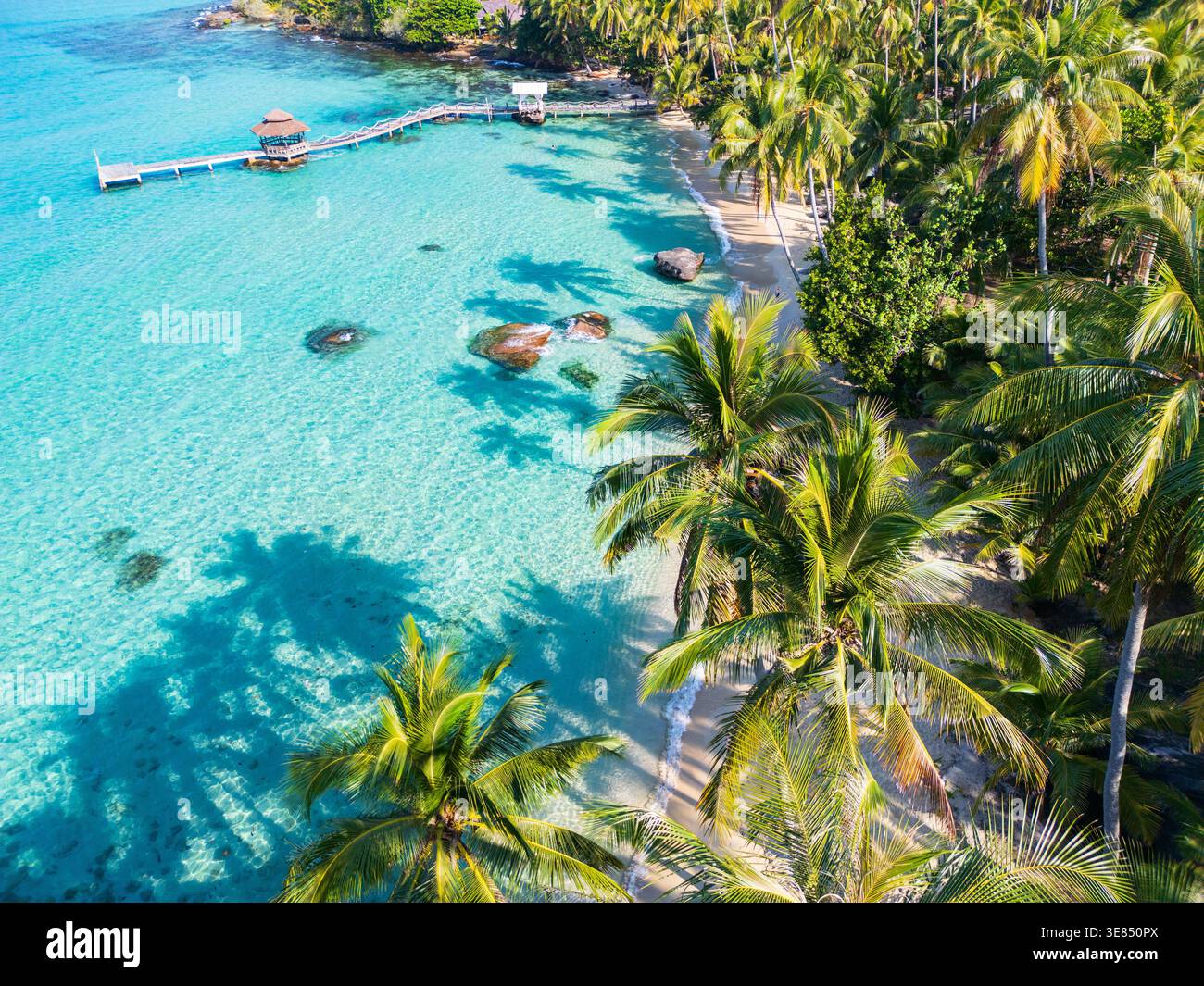 Blick aus der Vogelperspektive auf den tropischen Strand mit türkisfarbener Lagune, Palmen, weißem Sand und kleinem Pier, sonniges Inselurlaubsziel. Stockfoto