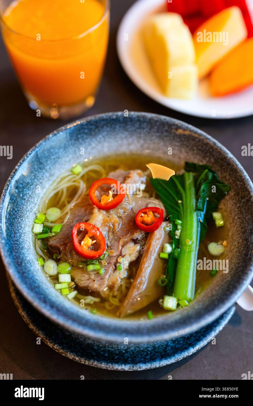 Schüssel asiatische Nudelsuppe mit geschmortem Rindfleisch, roten Chili-Scheiben, Zwiebeln und Pak Choy auf dunklem Tisch, mit Orangensaft und Obstteller im Hintergrund Stockfoto