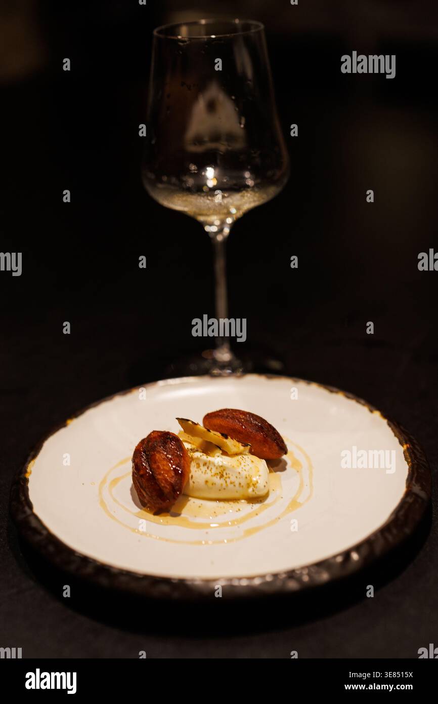 Feines Dining Dessertteller mit Sahnequenelle, karamelisierten Fruchtscheiben und Sauce Nieseln, mit leerem Weinglas im Hintergrund, stimmungsvollem, schwachem Licht, Sch Stockfoto