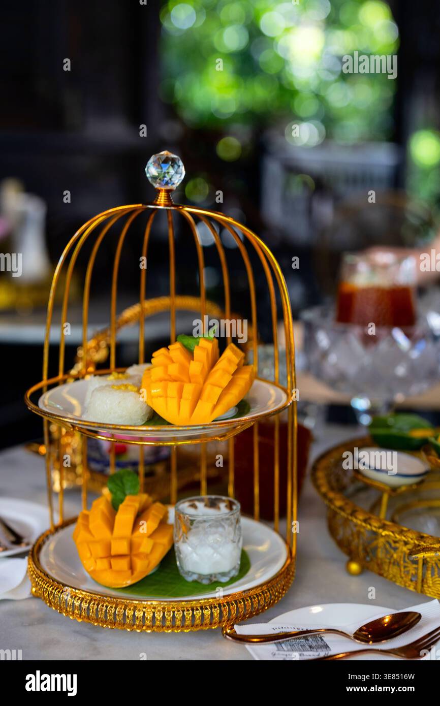 Luxuriöser Nachmittagstee mit Mangodessert, klebrigem Reis und Eis, serviert auf kunstvoll verzierten Goldtablett unter dekorativem Käfig, geringe Tiefe des Feldes, gehobene Klasse Stockfoto