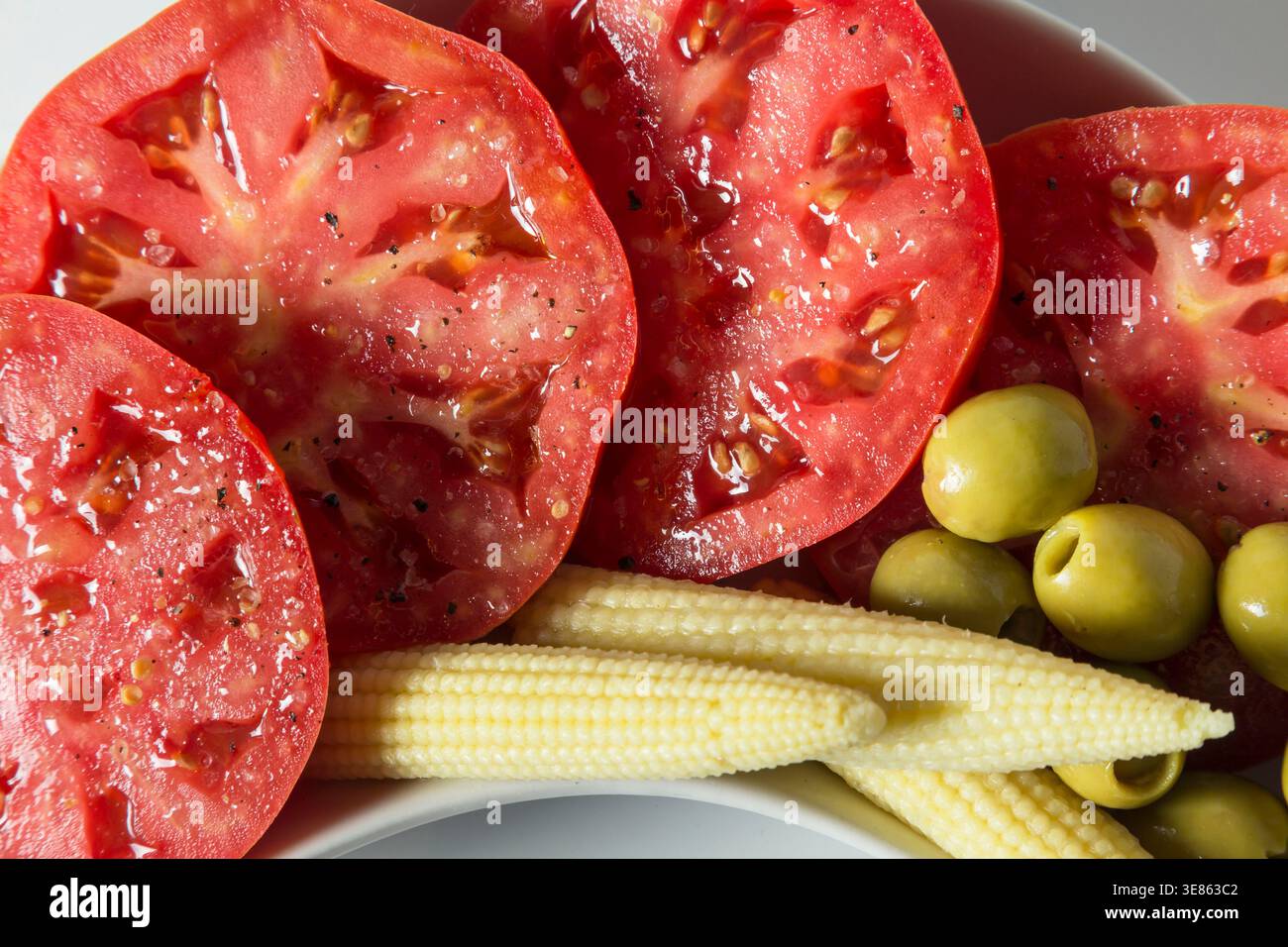 Eine hochschwingende Nahaufnahme saftiger Tomatenscheiben mit Salz und Pfeffer, serviert mit eingelegtem Babymais und grünen Oliven. Helles, natürliches Licht h Stockfoto