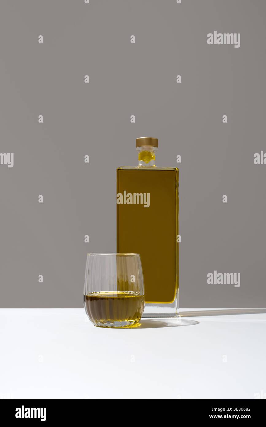 Minimales Stillleben mit einer quadratischen Flasche goldenem Olivenöl und einem kleinen Verkostungsglas auf einer weißen Oberfläche, weichem Licht und neutralem Hintergrund, Elegane Stockfoto