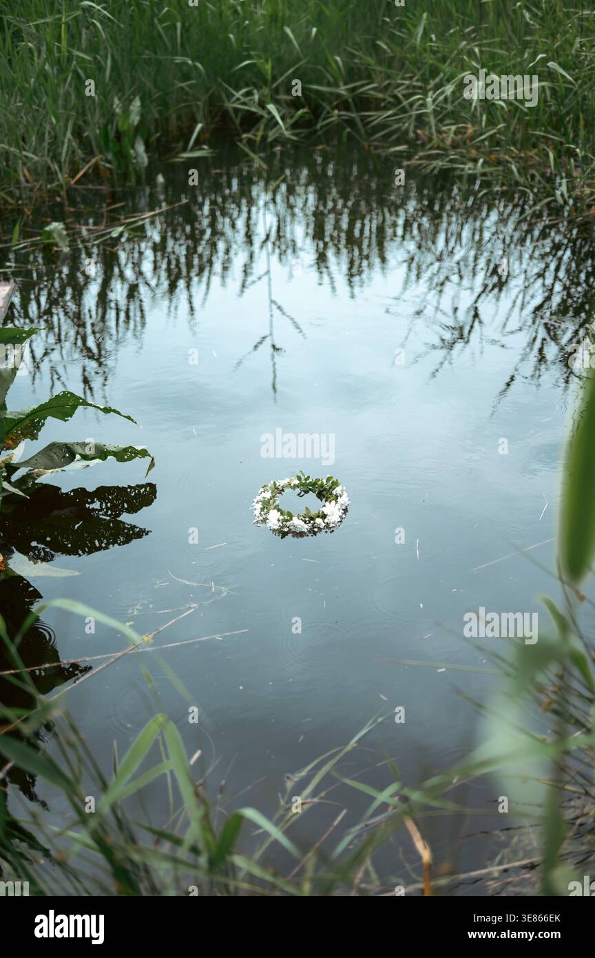 Ein zarter Wildblumenkranz schwimmt auf einem ruhigen, ländlichen Teich, umgeben von Schilf und Wiesengräsern. Eine heidnische Sommersonnenwende ruft rustikales Leben hervor, Stockfoto