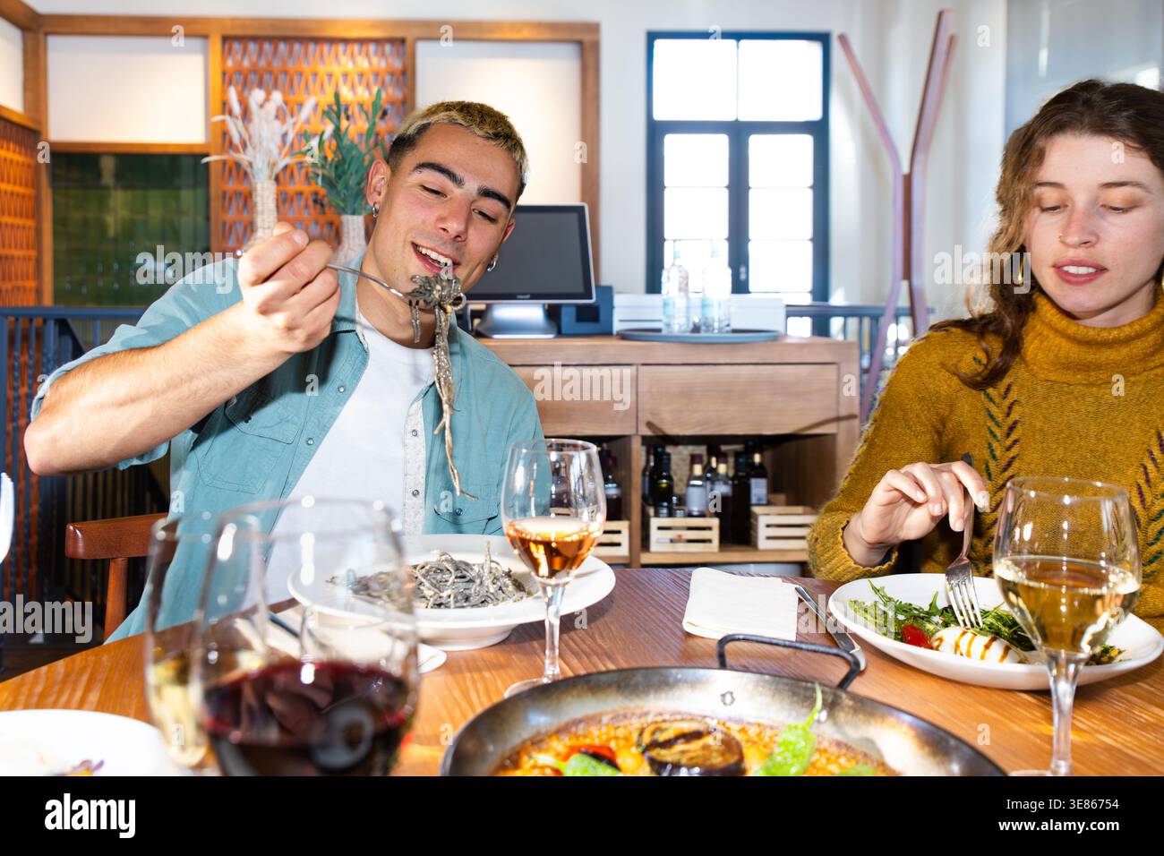 Freunde genießen eine mediterrane Mahlzeit in einem gemütlichen Restaurant und genießen Paella und Wein an einem Holztisch. Ein Mann serviert Nudeln, während eine Frau Salat isst Stockfoto