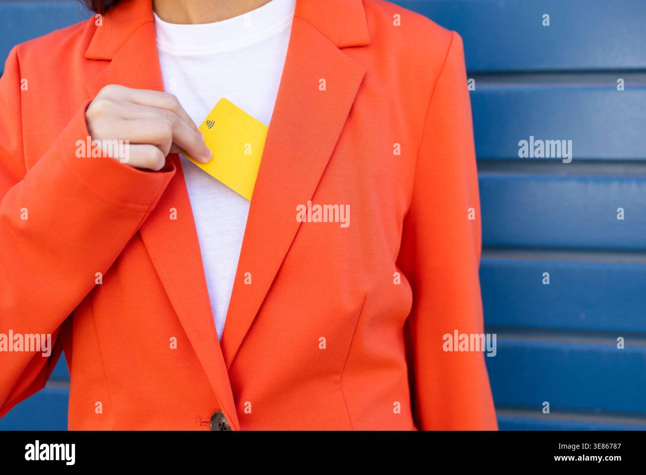 Eine Geschäftsfrau in einem leuchtend orangefarbenen Blazer steckt eine gelbe Kreditkarte in ihren Blazer und symbolisiert Finanztransaktionen und Geschäftsgeschäfte Stockfoto