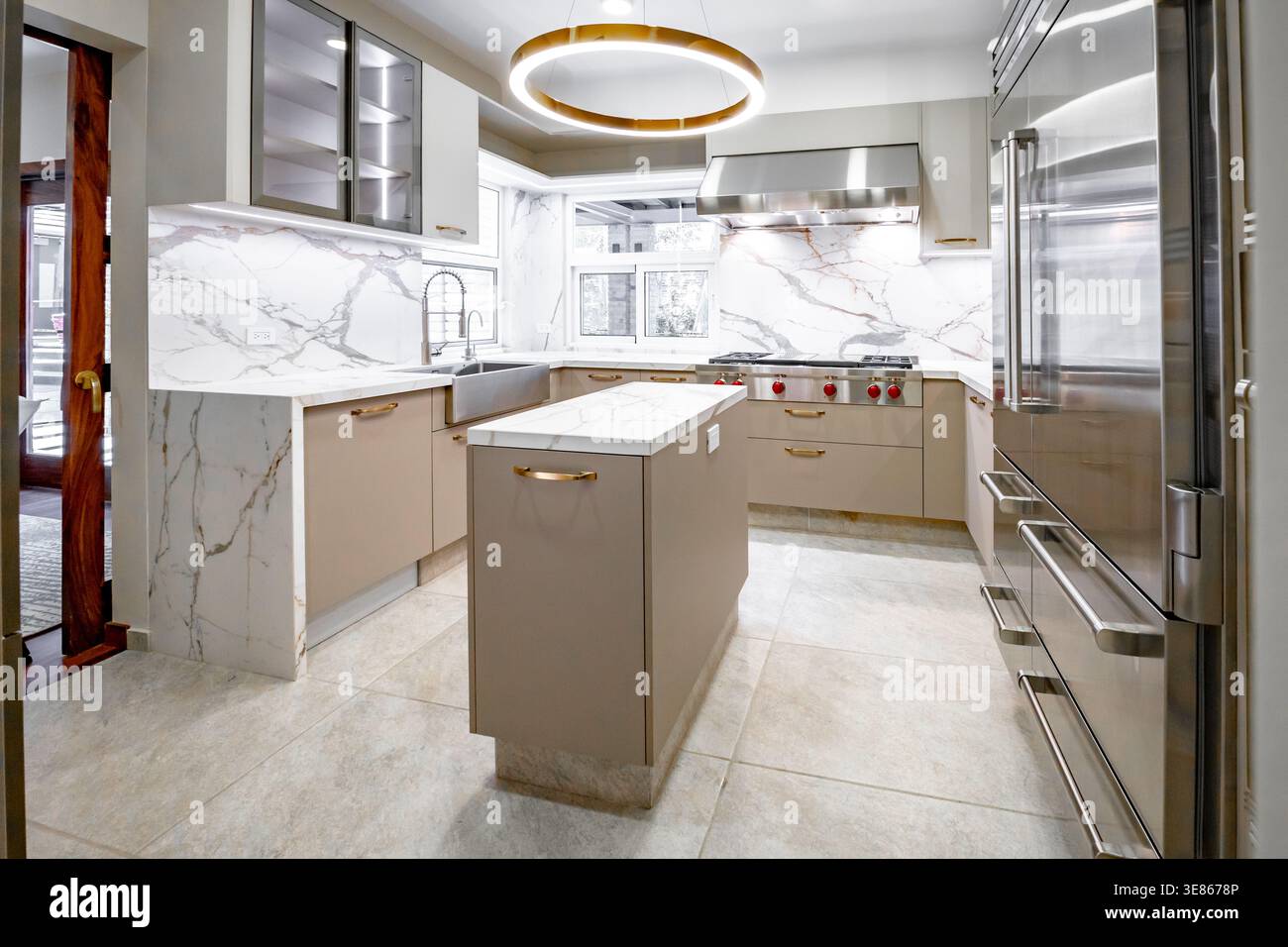 Helle, luxuriöse, minimalistische Kücheneinrichtung mit Marmorarbeitsplatten und Backsplash, eleganten Schränken, Edelstahlgeräten und zentraler Inselgruppe Stockfoto