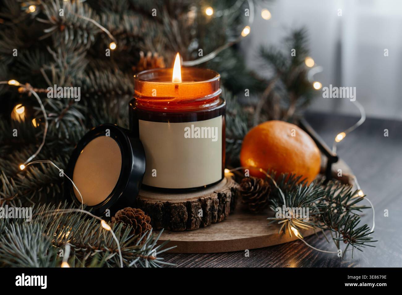 Eine gemütliche Szene mit einer beleuchteten Sojabauwachskerze mit einem leeren Etikett, umgeben von festlichen Dekorationen wie Kiefernzweigen, einer Mandarine und Weihnachtslichtern Stockfoto