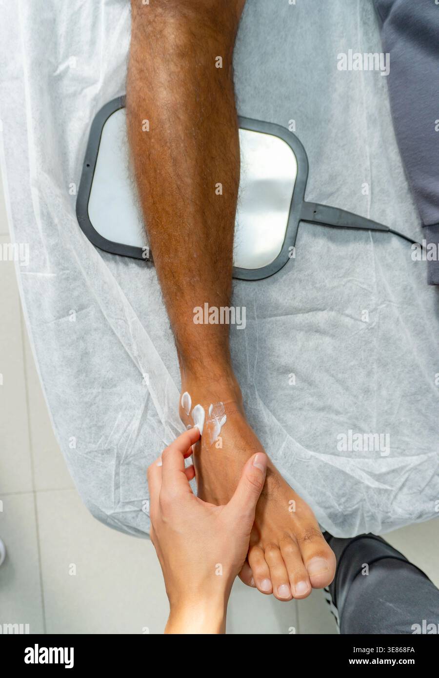 Oben Nahaufnahme einer Hand eines Therapeuten, die vor der Behandlung mit Rehabilitationsgeräten Creme auf den Knöchel des Patienten aufträgt, wobei Hautpflege und Schmerzreaktionen hervorgehoben werden Stockfoto