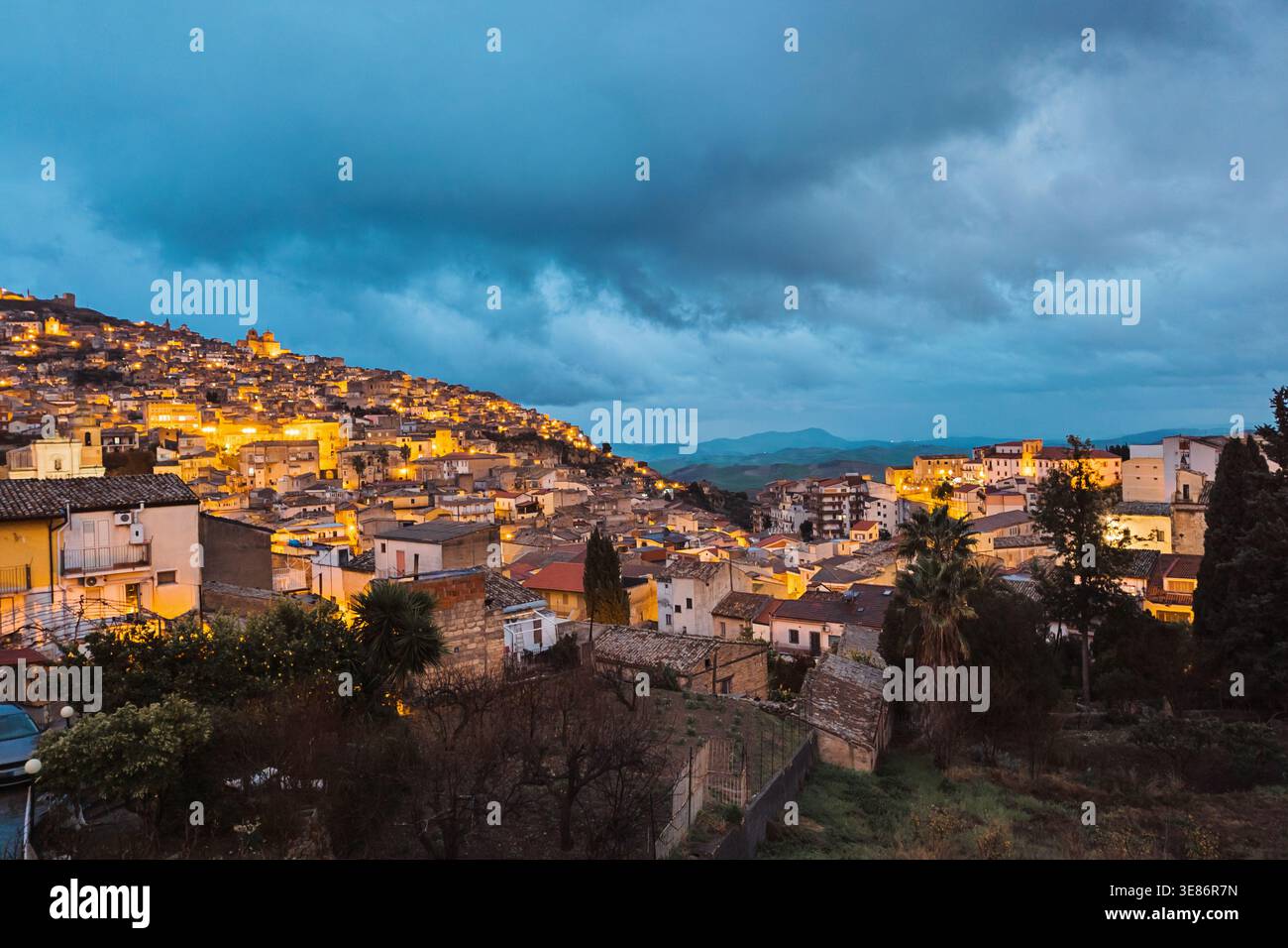Hill Town in der Abenddämmerung mit leuchtenden Lichtern unter dramatischen Sturmwolken Stockfoto