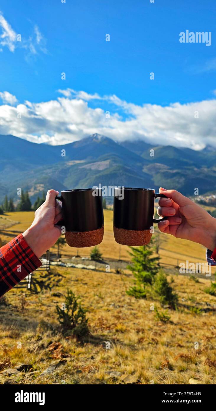 Kaffeetassen Cheers mit Bergblick. Zwei Hände halten schwarze Kaffeetassen vor einer malerischen Berglandschaft und genießen einen ruhigen Morgen im Freien Stockfoto