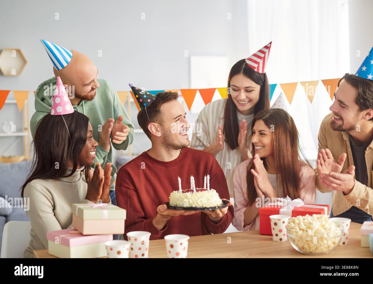 Fröhliche Gruppe von verschiedenen Freunden mit Partyhüten, die zu Hause mit Kuchen und Geschenken ihren Geburtstag feiern. Stockfoto