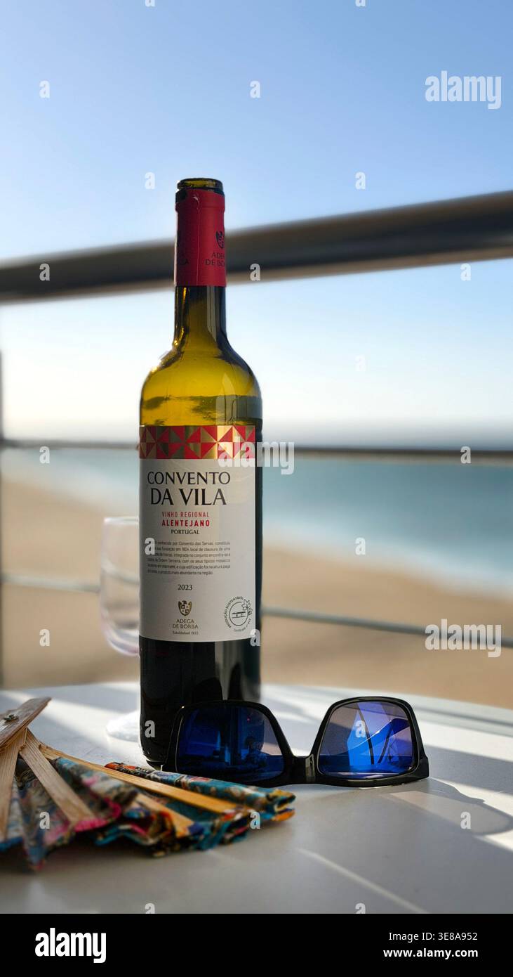 Flasche Rotwein Convento da Vila Alentejano auf einem Tisch mit Sonnenbrille und Ventilator mit Blick auf einen sonnigen Strand in Portugal Stockfoto