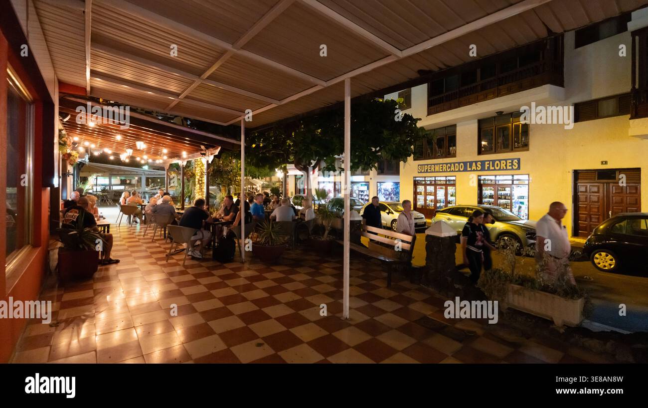 Eine lebhafte Abendszene entfaltet sich, in der die Gäste ihre Mahlzeiten an Tischen auf einer karierten Terrasse genießen, die von warmen Lichtern beleuchtet wird, und in einem nahe gelegenen Supermarkt Stockfoto