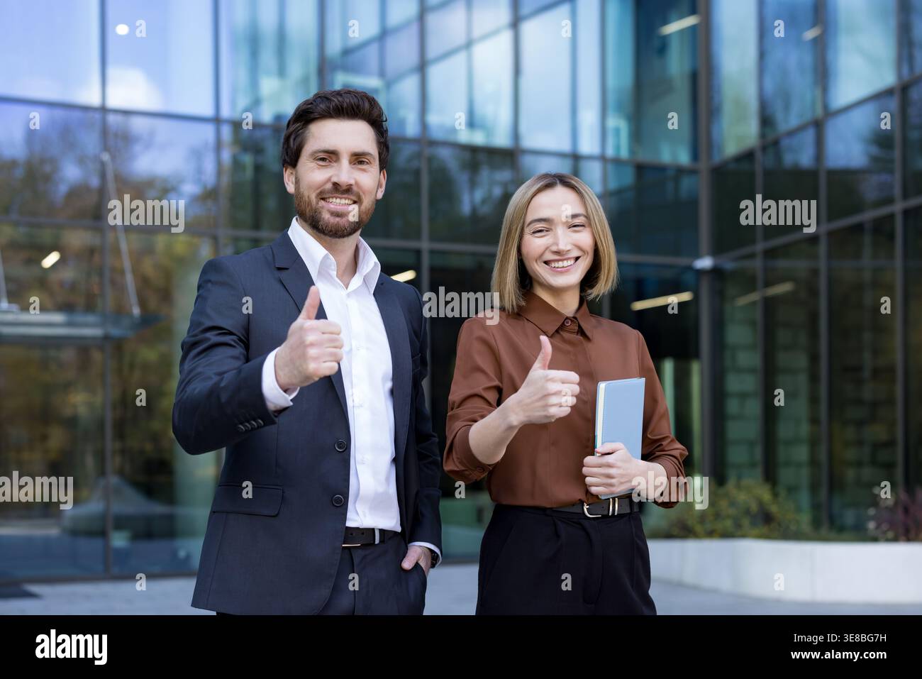 Zwei lächelnde Geschäftskollegen, ein Mann und eine Frau, die vor einem modernen Bürogebäude stehen und beide eine Geste nach oben geben, die Erfolg, Zusammenarbeit und Anerkennung repräsentieren Stockfoto