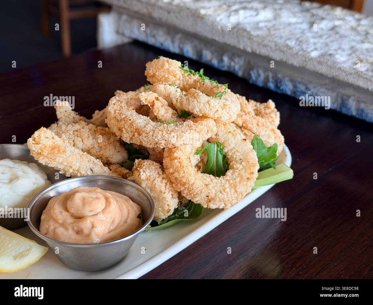 Knusprige goldene gebratene Calamari-Ringe mit Dippsoße auf einem weißen Teller, eine klassische Vorspeise im Restaurant. Saubere Lebensmittelfotografie in warmen Tönen, ideal für Menügestaltung, Speisen und kulinarische Lifestyle-Inhalte. Stockfoto