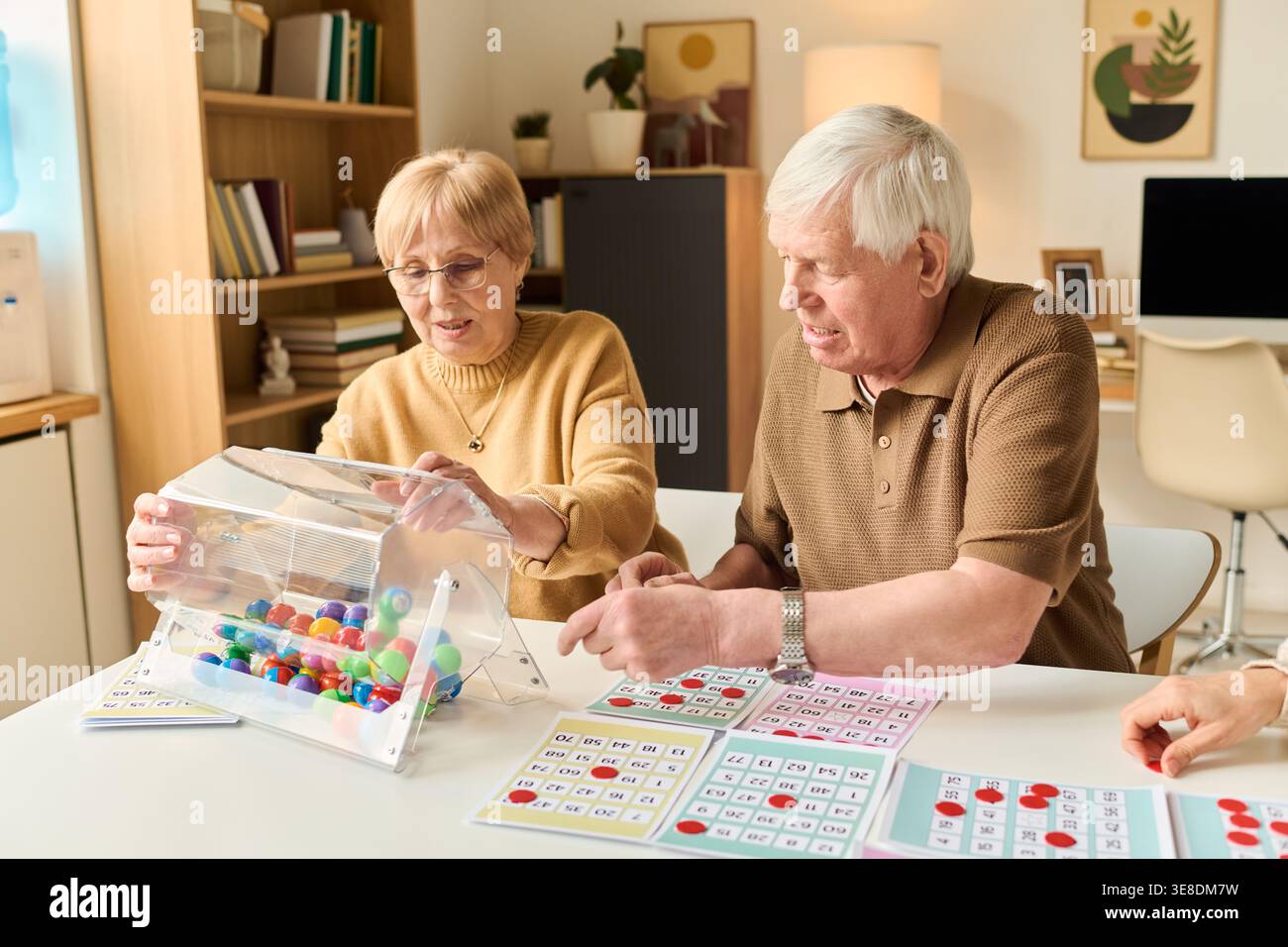 Ältere kaukasische Ehepaare spielen am Tisch Bingo, nehmen kognitive Aktivität und soziale Interaktion wahr. Nützlich für den Ruhestand-Lebensstil, Gedächtnispflege, Senioren Wellness-Marketing Stockfoto