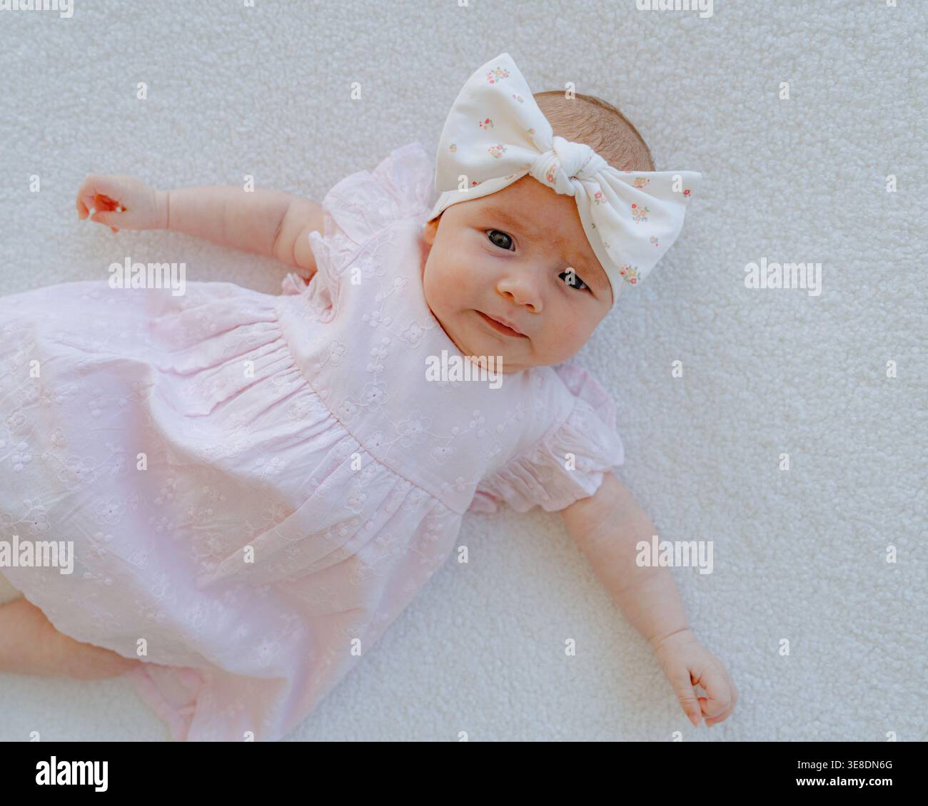 Niedliches Mädchen mit rosa Kleid und Schleife, das auf weichem weißem Hintergrund liegt. Minimales und ästhetisches Babyporträt mit Kopierraum. Erziehung und Kindheit Stockfoto