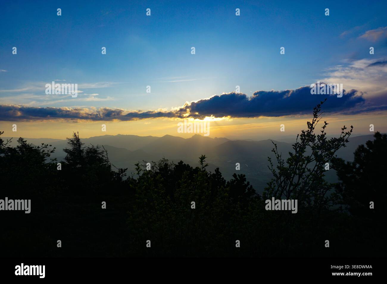 Sonnenuntergang hinter einer Bergkette mit goldenem Licht, das durch Wolken über ein üppiges Tal strömt. Stockfoto