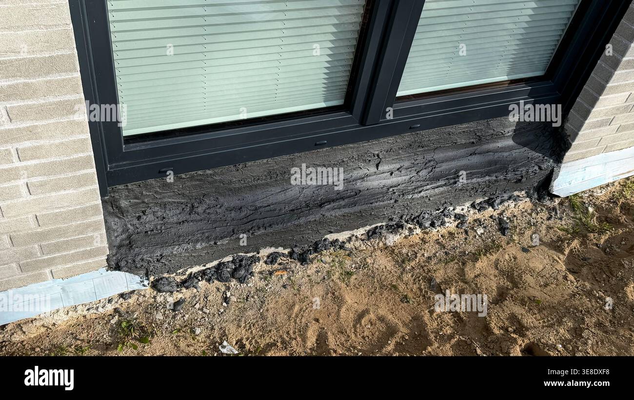 Nahaufnahme eines frisch aufgetragenen schwarzen Dichtmittels um die Basis eines modernen Fensters in einem Wohngebäude mit sichtbarem Sandboden und Ziegelwand Stockfoto