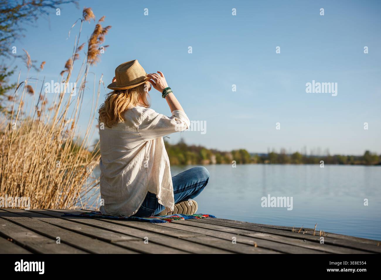 Frau mit Hut, die vom Dock aus über den friedlichen See blickt und Sonne und frische Luft im Freien genießt. Einsame Entspannung und Achtsamkeit in der Natur Stockfoto