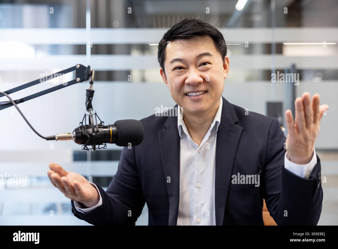 Asiatischer Geschäftsmann, der selbstbewusst Gesten in ein professionelles Studio-Mikrofon einnimmt und ansprechende Audioinhalte für einen Podcast oder ein Online-Interview erstellt Stockfoto