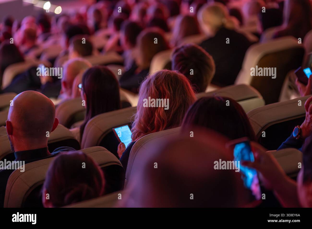 Während der Konferenz saßen die Teilnehmer in einem dimmen Auditorium mit Smartphones. Die warme Bühnenbeleuchtung sorgt für intime Stimmung. Stockfoto