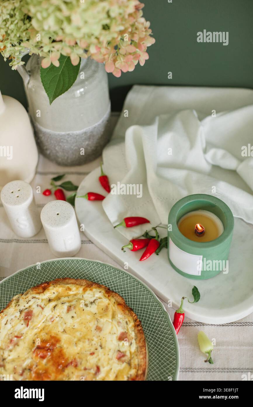 Blick von oben auf eine herzhafte Quiche auf einem Tisch mit einer duftenden Kerze, einen Haufen frischer Hortensie-Blumen in Vase, Salz- und Pfefferstreuer und rote Chilischoten Stockfoto