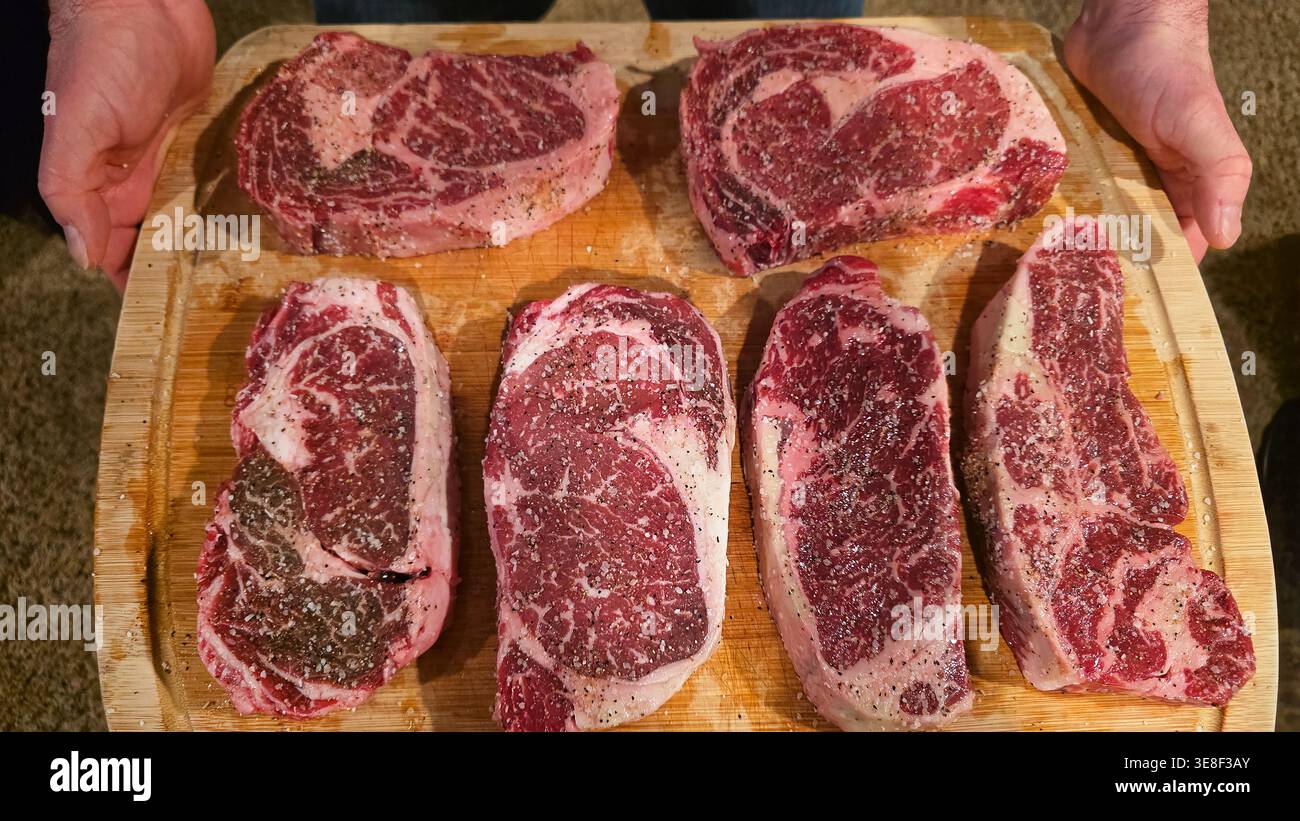 Rohe Ribeye-Steaks, gewürzt auf Holzbrett. Nahaufnahme mehrerer roher marmorierter Ribeye-Steaks, die mit Salz und Pfeffer auf einem hölzernen Schneidebrett gewürzt wurden Stockfoto
